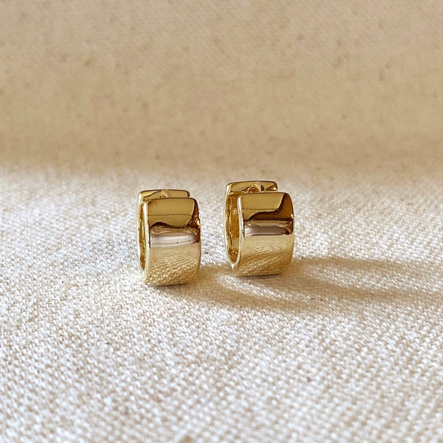 A pair of gold chunky flat clicker hoop earrings placed on a beige textile surface.