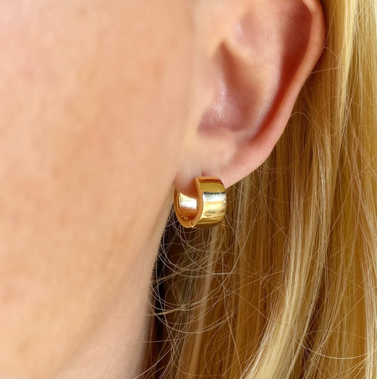 A pair of gold chunky flat clicker hoop earrings placed on a beige textile surface.